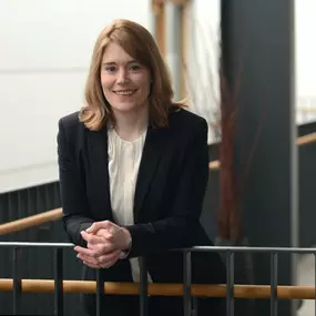 Frau im Blazer lehnt am Geländer in einem modernen Atrium; weiches Licht, unscharfer Hintergrund, freundlicher Blick.