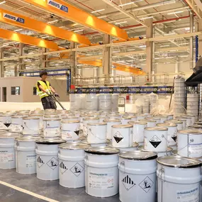 Viele Eimer mit Gefahrstoffsymbolen in Fabrikhalle; zwei Arbeiter zwischen Gebinden, gelbe Deckenkrane im Hintergrund.