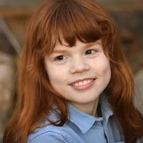 Kinderportrait draußen: rothaariges Kind mit Sommersprossen im blauen Hemd, schaut seitlich; Hintergrund unscharf.
