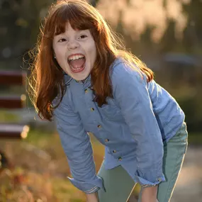 Rothaariges Kind lacht nach vorn gebeugt im Abendlicht; deutliche Lichtkante, Hintergrund weich unscharf.