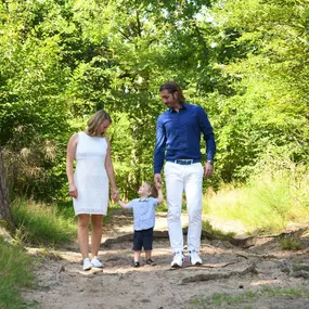 Familie mit kleinem Kind geht Hand in Hand auf einem sandigen Waldweg; grüne Bäume, zwei Stämme rahmen die Szene.