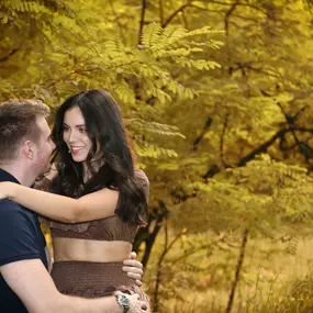 Paar in herbstlicher Natur, sie in seinen Armen; weiches Vorderlicht, warmes Gegenlicht, Hintergrund stark unscharf.