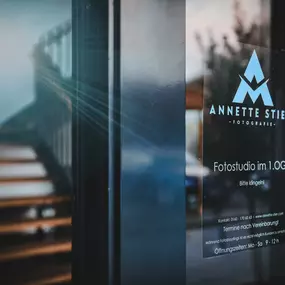Offene Eingangstür mit Firmenschild rechts auf Glas („Annette Stier – Fotostudio im 1. OG – bitte klingeln“), Treppe im Hintergrund.