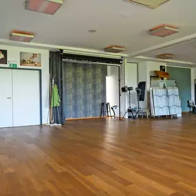 Innenraum des Fotostudios aus weiterer Perspektive: große freie Fläche mit Holzfußboden, Deckenleuchten, Hintergrundsystemen, Stativen und Requisitenwagen. Ideal für variable Sets von Business- bis Familien-Shootings bei ‚ANNETTE STIER – Fotografie‘.