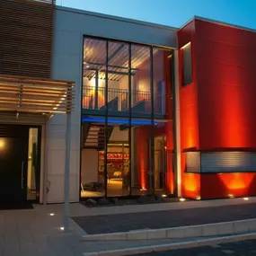 Moderne Studiofassade in Bensheim: rote Paneelwand, große Glasfront, Holzlamell-Vordach und Bodenleuchten bei Dämmerung.