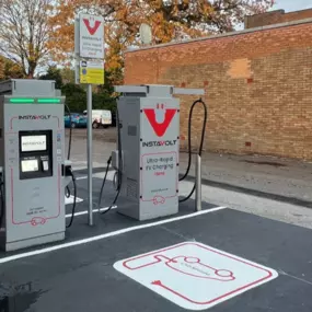 Two InstaVolt ultra-rapid chargers located in a lined charging bay next to a brick wall, with autumn trees visible in the background and marked EV symbols on the ground.
