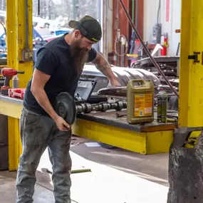 A bearded mechanic in a workshop holds a gear while inspecting engine parts, including a camshaft, on a yellow workbench near a large oil container.