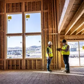 Two construction professionals wearing safety vests and hard hats stand inside a partially built house, reviewing plans on a tablet. The unfinished interior features exposed wooden framing and large installed windows that offer a clear view of a residential development and hills in the distance. Natural light fills the room, highlighting the clean structure and organized construction site outside.