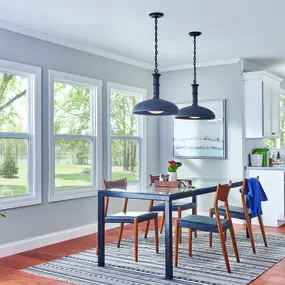 Bright daylight streams into an open-concept dining room through a trio of prominent white double-hung windows, showcasing a well-maintained backyard with mature trees.