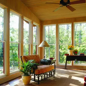 A sunroom featuring a full wall of tall, white-framed double-hung windows, a textured sofa, a small wooden table with flowers, and a ceiling fan, all overlooking a vibrant woodland.
