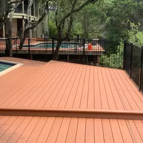 A spacious, terracotta-colored composite deck built adjacent to an in-ground swimming pool. The modern design features black aluminum railings, multiple levels, and overlooks a lush, green wooded area.