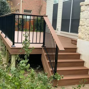 A raised composite deck featuring brown planks and black aluminum railings attached to a cream house with stone accents. A staircase leads down to a garden area with lush green foliage underneath.