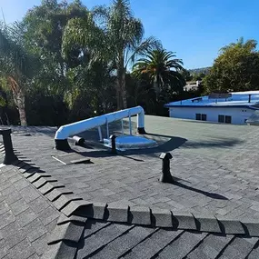 A residential roof with brown and black architectural asphalt shingles features several plumbing vent pipes and a transition to a flat, red granule-surfaced roof section.