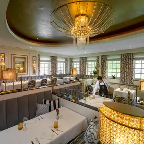 Das stilvolle Orangerie-Restaurant präsentiert sich mit großen Glasfenstern, durch die natürliches Licht den eleganten Speisesaal erhellt. Im Vordergrund stehen fein gedeckte Tische mit weißem Porzellan und frischen Blumenarrangements.