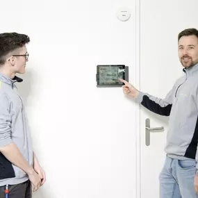 Zwei unserer Gebäudeinformatiker die vor einem Touchpanel für die Smarthome-Anwendung stehen. Sie sind zuständig für Arbeiten rund um Energieeffizienz, intelligente Schaltanlagen und Multimedia-Systeme.