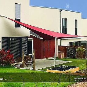 rotes, gespanntes Sunfurl-Sonnensegel, in einem kleinen, grünen Garten mit modernem, weißem Haus.