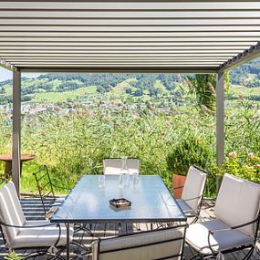 halb-transparente, weiße Standmarkise über einer Terrasse mit Glas-Tisch und Stühlen, inmitten der Natur.