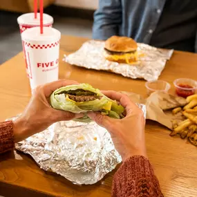 A customer's hands hold a Five Guys lettuce wrap at a dining room table inside a Five Guys restaurant.