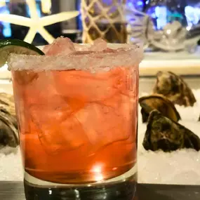 A refreshing cocktail served at Slacks Oyster House, Plainridge Park Casino, placed in front of a bed of fresh oysters on ice. The drink is garnished with a lime wedge, making it the perfect pairing with the restaurant's seafood offerings.