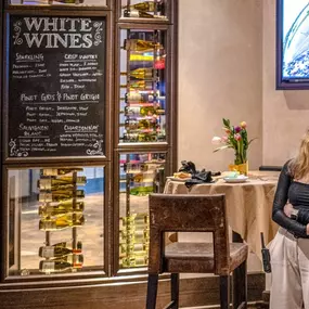 The wine selection area at Slacks Oyster House, Plainridge Park Casino, featuring a floor-to-ceiling display of fine wines. A chalkboard highlights the restaurant's offerings of white wines, including Chardonnay and Pinot Grigio, adding to the upscale ambiance.