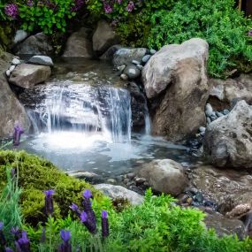 A Greenhaven Landscapes-designed water feature - a Vancouver WA backyard waterfall with large stones and pebbles along the bank and shrubbery running up the hillside