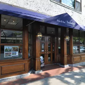 White brick building with dark wood storefront and blue awning housing Julia B. Fee Sotheby's International Realty in Bronxville, New York, featuring property listing displays in windows.